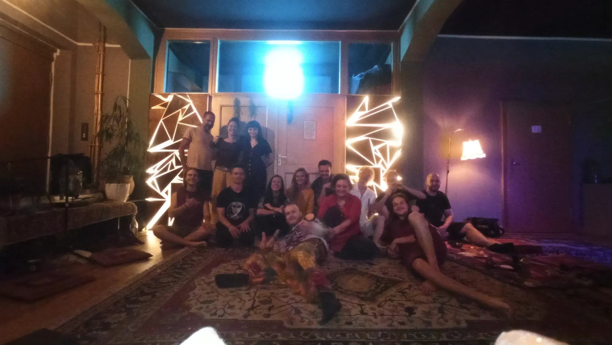 Group photo after a cacao ceremony in a studio, people sitting and lying on a rug in front of glowing triangle lights