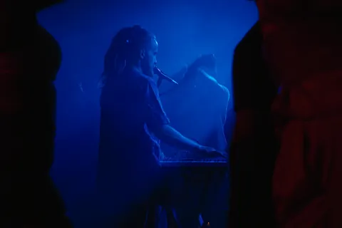 Gray Contrast singing into a microphone at his keys in blue stage haze, dancers in silhouette in the foreground